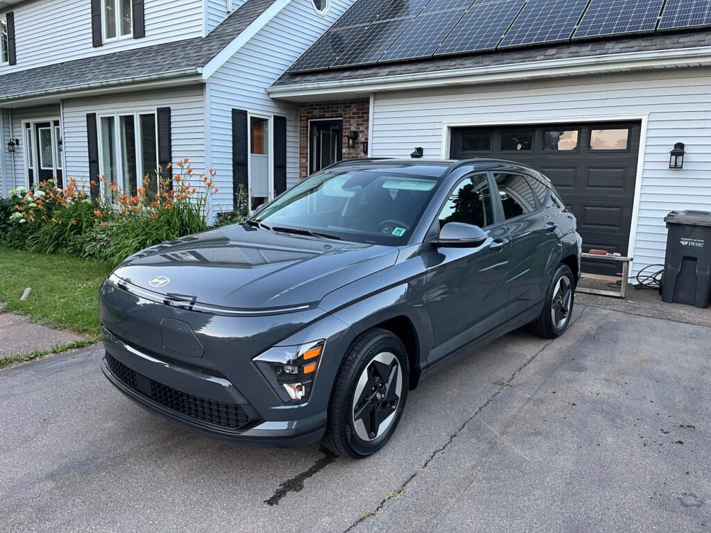 Grey Hyundai Kona EV in driveway in front of a suburban home with solar panels and flower beds.