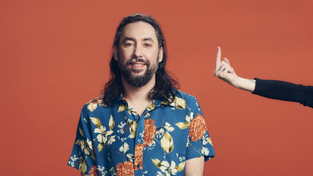 Brad wearing a floral-pattern shirt with long pandemic hair and a hand giving him the middle-finger.