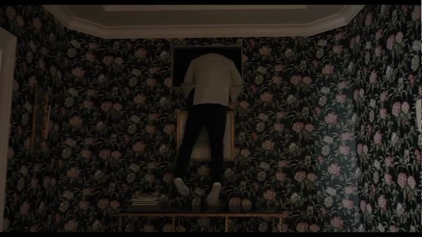 Frame from the show Only Murders in the Building with a person climbing out of an air duct into a room with dark floral wallpaper.
