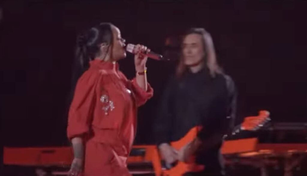 Rihanna wearing red singing into a microphone in front of a man with a red guitar