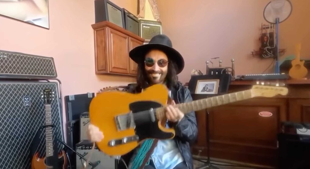 Still from video of Mike Campbell smiling and wearing a had and sunglasses, holding a Broadcaster guitar in front of some guitar amps.