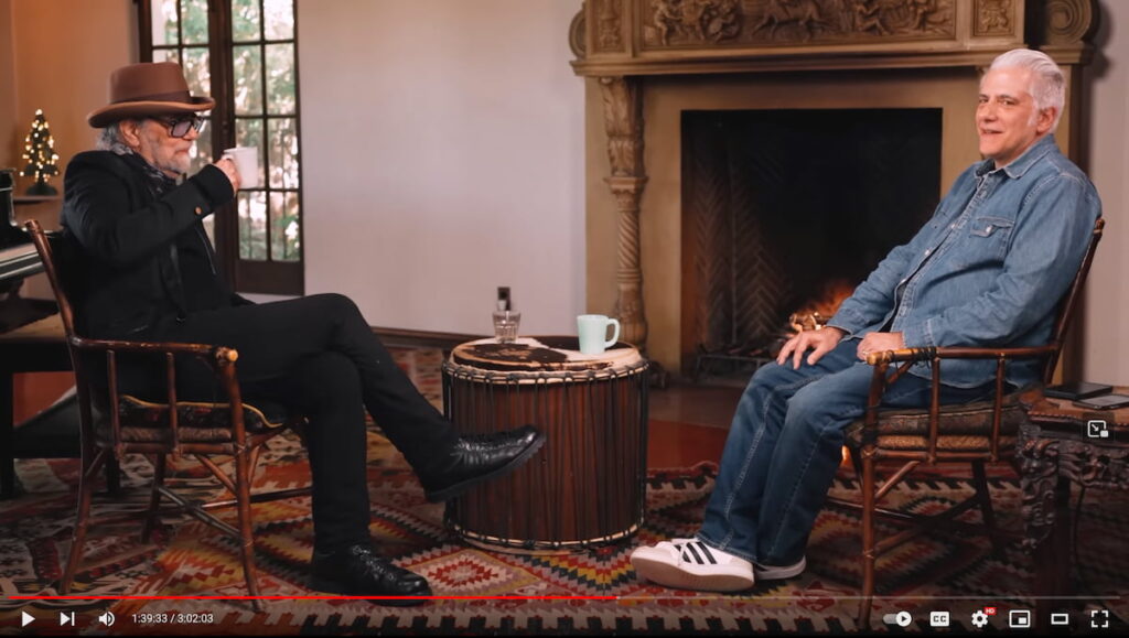 Screenshot from YouTube interview showing Daniel Lanois in a hat and sunglasses drinking from a mug across from Rick Beato who is all in denim, in a comfortable living room setting.
