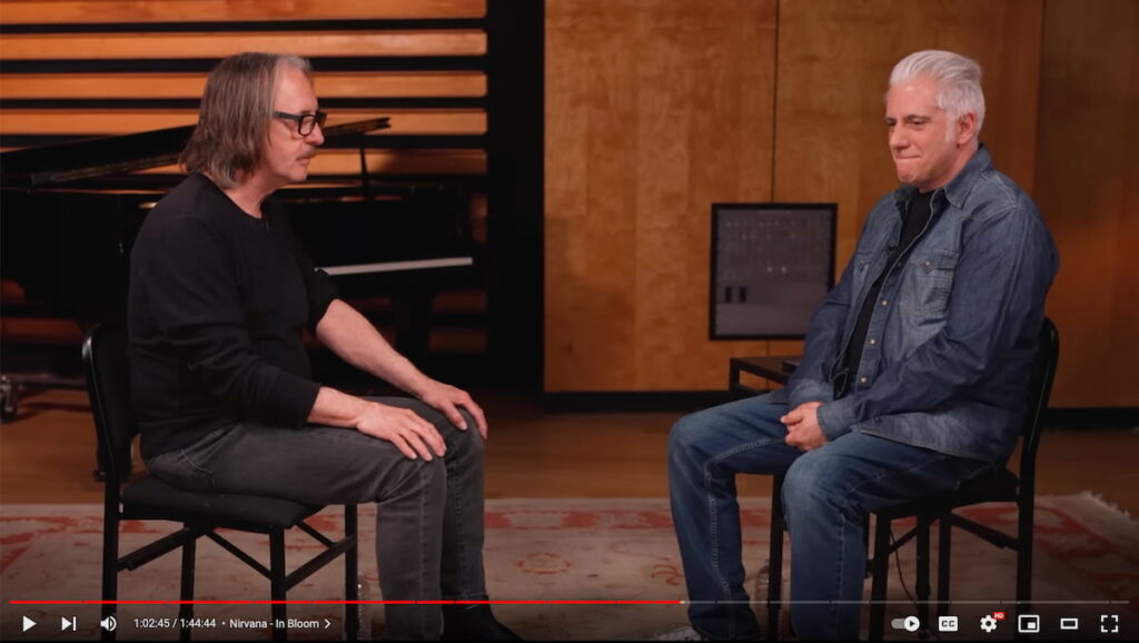 Screenshot from a YouTube video with Butch Vig with long hair and heavy-framed glasses sitting across from Rick Beato all in demin in a music studio setting
