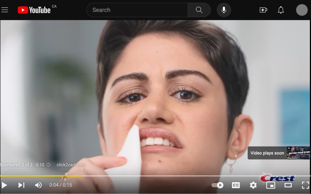 Screenshot of YouTube website with still image from video ad showing a woman holding a while kleenex up by her teeth to compare the shade of white, and making a displeased face.